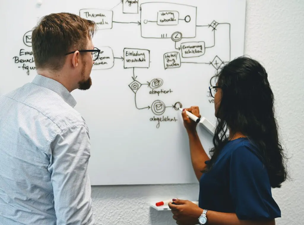 Zwei junge Menschen, die auf ein bemaltes White Board blicken und mit einem schwarzen Stift ein Element ergänzen.