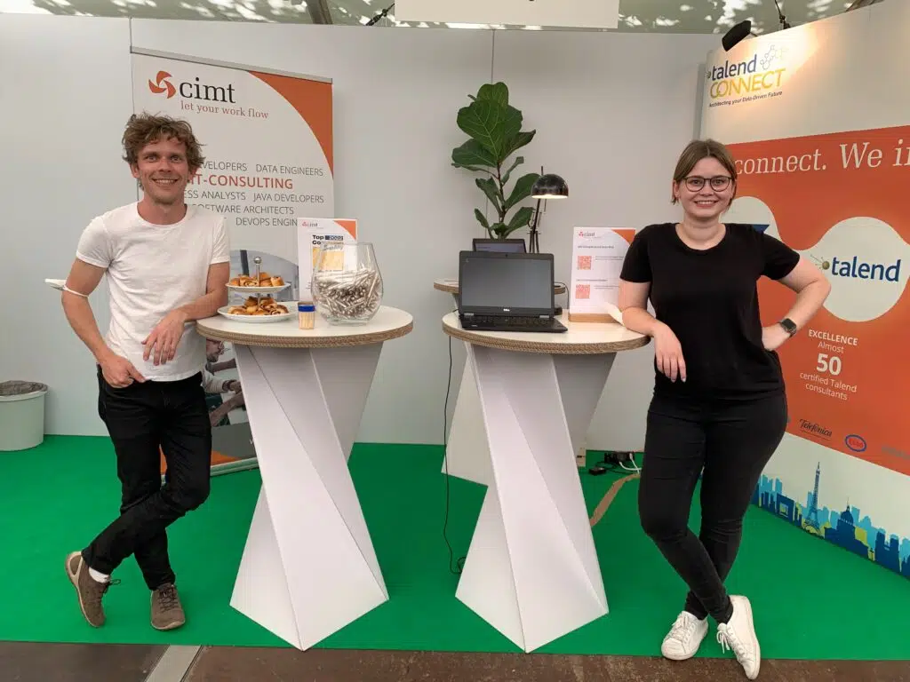 Zwei Personen stehen am Messestand und lehnen sich an den Tisch an.