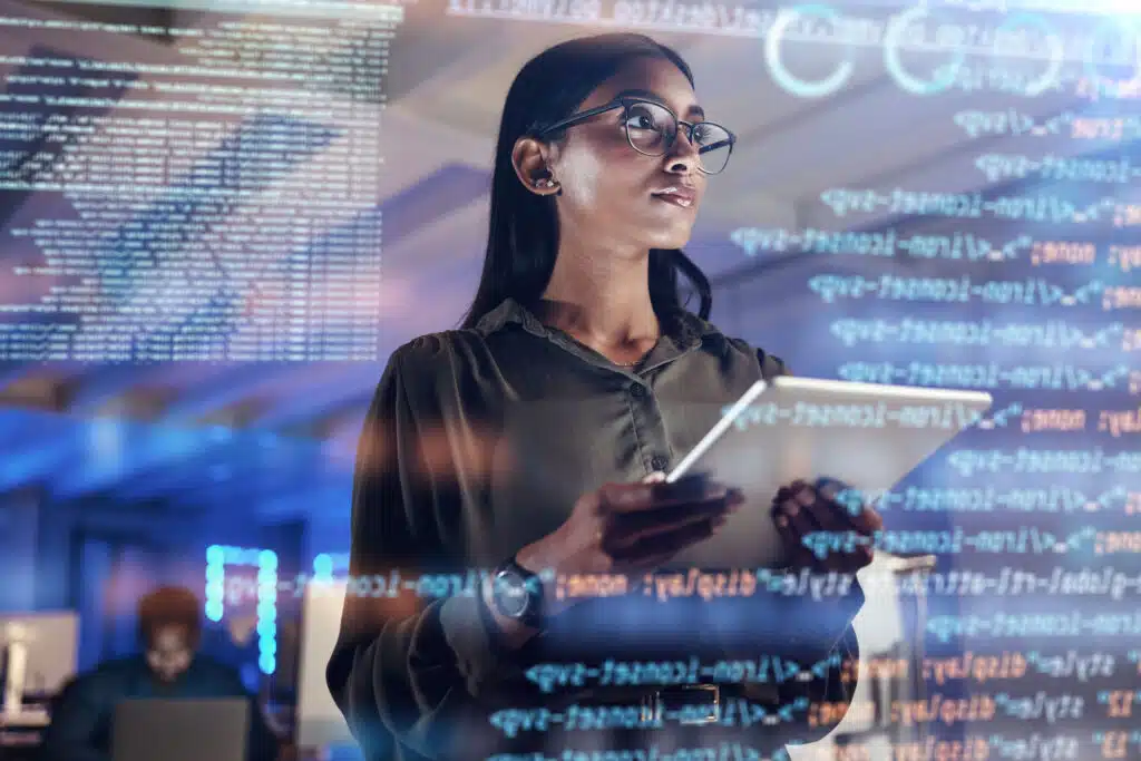 Eine Frau blickt nach oben und hält ein Tablet in der Hand. Im Vordergrund ist eine transparente Wand mit Daten zu sehen.